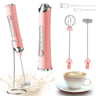 Pink electric milk frother with attachments and charging cable, beside cup of coffee.