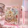 Pink notebook with 3D miniature scenes and stickers, standing on a desk with a fan, lamp, and other stationery items.